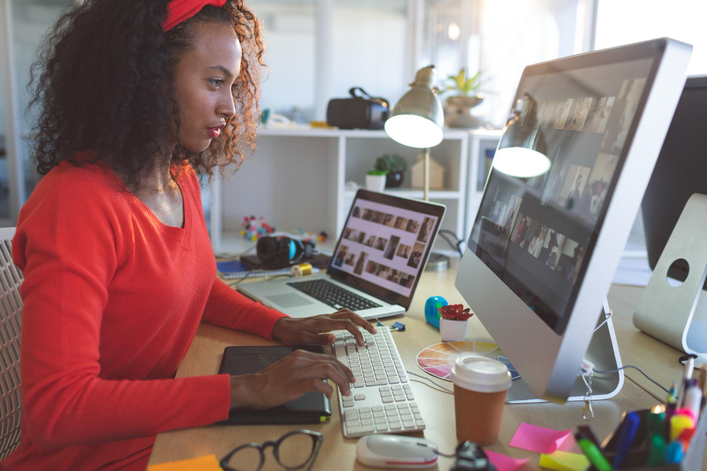 Smiling South African woman, working at a modern desk setup with dual monitors, fully focused on designing her brand’s online store, surrounded by creative tools and a warm, professional workspace.